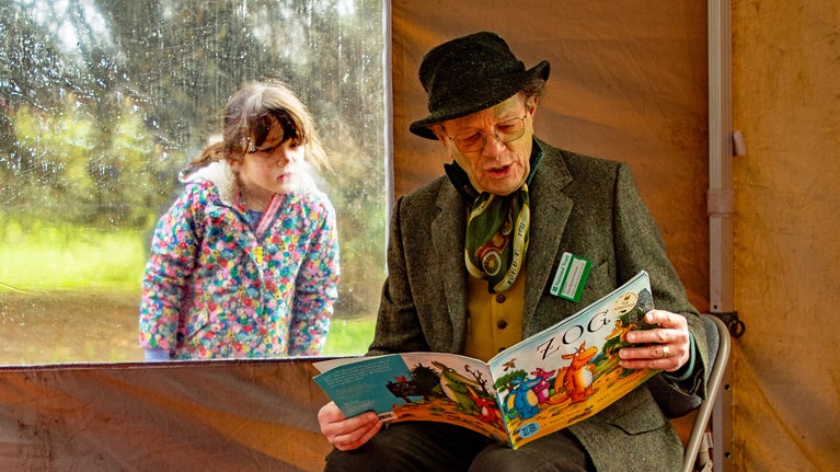 A man wearing a bowler hand and waistcoat reads the picture book "Zog" while sitting in a tent. A young girl peers in through the tent window to watch him.